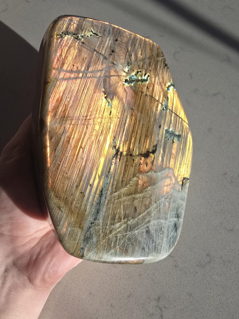 Puede incluir: Una losa de piedra labradorita pulida con una forma rectangular redondeada y lisa. La superficie muestra una vibrante iridiscencia dorada y bronce con estr&iacute;as lineales visibles e inclusiones minerales oscuras y dentadas.