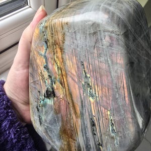 Puede incluir: Una piedra de labradorita pulida con una superficie lisa y redondeada con fracturas y capas internas visibles. La piedra muestra destellos iridiscentes de oro, rosa y azul sobre una base gris oscuro semitransl&uacute;cida con patrones lineales.