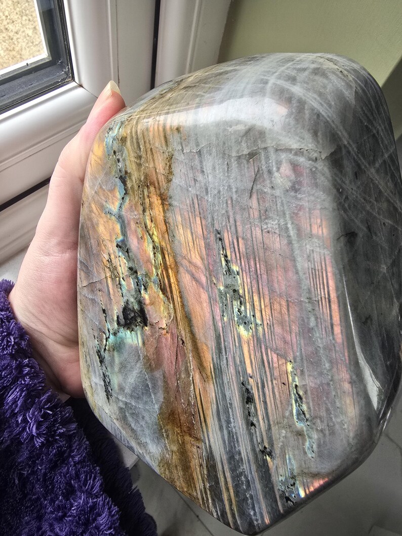Puede incluir: Una piedra de labradorita pulida con una superficie lisa y redondeada con fracturas y capas internas visibles. La piedra muestra destellos iridiscentes de oro, rosa y azul sobre una base gris oscuro semitransl&uacute;cida con patrones lineales.