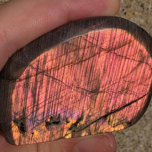May include: A polished labradorite stone with a dark gray base and a prominent, vibrant flash of iridescent pink, orange, and gold light across its surface. The stone features visible linear striations and natural fissures throughout its structure.