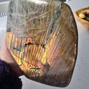 Puede incluir: Una piedra de labradorita pulida de forma irregular con una superficie lisa y fracturas y estr&iacute;as internas visibles. La piedra muestra un destello dorado-anaranjado prominente en el centro, que transiciona a tonos grises y marrones.
