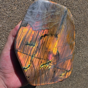 Puede incluir: Una piedra de labradorita pulida de forma irregular que presenta un vibrante despliegue de destellos iridiscentes en naranja, oro y amarillo sobre un fondo gris oscuro fibroso, con fisuras naturales visibles y una superficie lisa y reflectante.