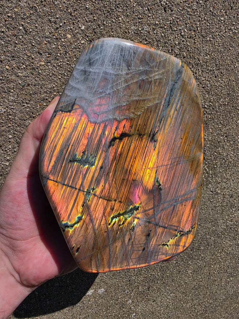 Puede incluir: Una piedra de labradorita pulida de forma irregular que presenta un vibrante despliegue de destellos iridiscentes en naranja, oro y amarillo sobre un fondo gris oscuro fibroso, con fisuras naturales visibles y una superficie lisa y reflectante.