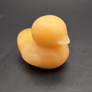 May include: A small, orange-yellow duck figurine. The duck has a smooth, rounded body and a small, pointed beak. The figurine is made of a solid material and has a slightly textured surface. The duck is a decorative item.