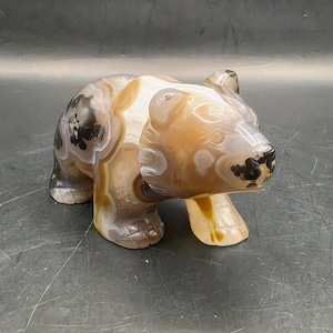May include: A carved bear figurine made of polished agate stone. The sculpture features natural banded patterns in shades of translucent brown, tan, cream, and white, with dark black inclusions on the snout and back, showcasing a smooth texture.
