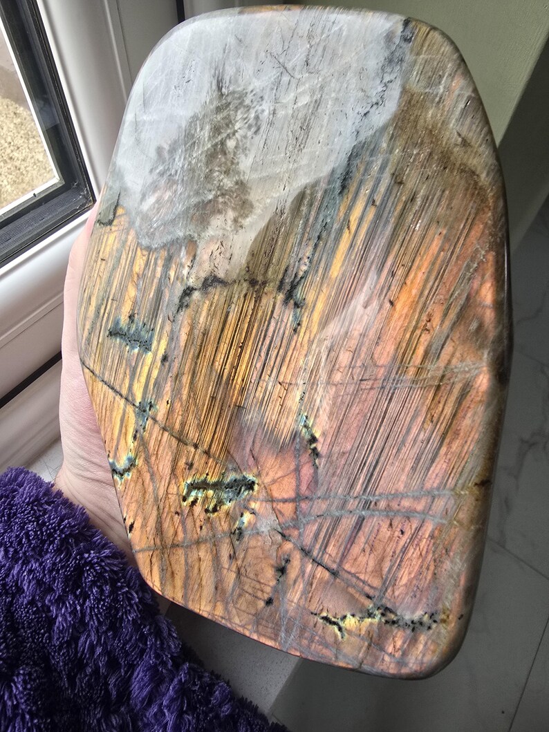 Puede incluir: Una losa de piedra labradorita pulida en forma de escudo, con una superficie lisa y prominentes estr&iacute;as lineales. La piedra muestra una paleta de colores &aacute;mbar c&aacute;lido, naranja dorado y marr&oacute;n oscuro, contrastada por una gran mancha blanca.