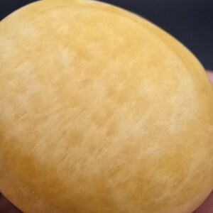 May include: A close-up of a smooth, oval-shaped object with a pale yellow color and subtle marbling. The surface has a slightly textured appearance. The object is set against a dark background.