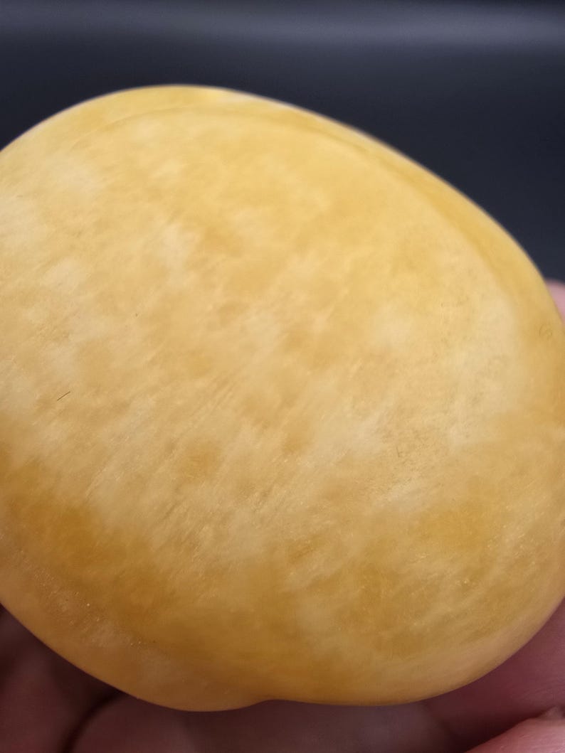 May include: A close-up of a smooth, oval-shaped object with a pale yellow color and subtle marbling. The surface has a slightly textured appearance. The object is set against a dark background.
