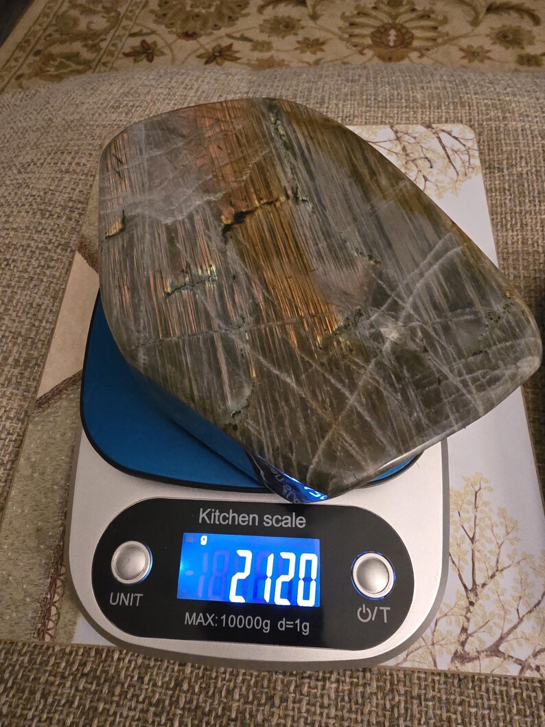 Puede incluir: Una piedra de labradorita pulida con vetas iridiscentes grises, marrones y doradas sobre una b&aacute;scula de cocina digital plateada y azul. La pantalla muestra un peso de 2120 gramos, con una capacidad m&aacute;xima de 10000g y una graduaci&oacute;n de 1g.