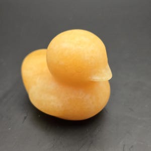 May include: A small, orange-yellow duck figurine with a smooth, rounded body and head. The duck has a small, pointed beak and is made of a solid, opaque material. The figurine is sitting on a dark surface.
