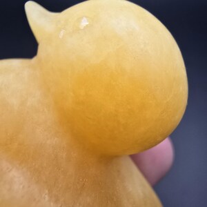May include: A close-up of a yellow duck figurine. The duck has a rounded body and head with a small beak. The figurine is made of a translucent material, and the color is a warm, golden yellow.