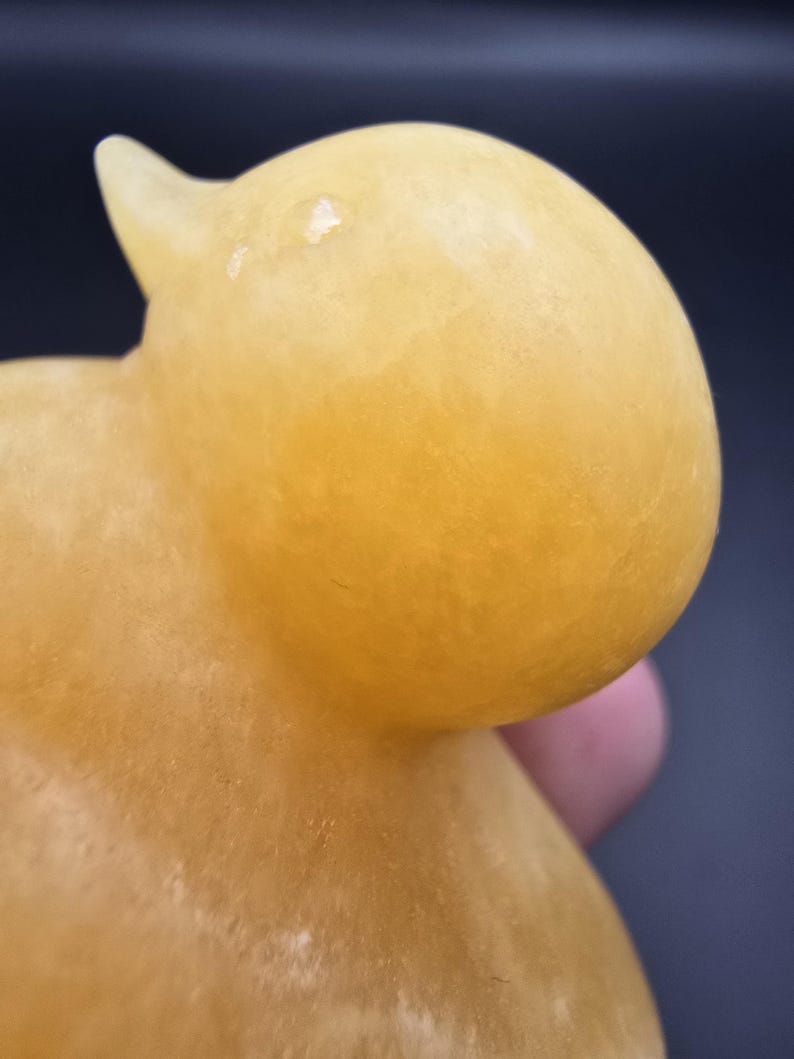 May include: A close-up of a yellow duck figurine. The duck has a rounded body and head with a small beak. The figurine is made of a translucent material, and the color is a warm, golden yellow.
