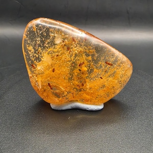 Peut inclure: Une pierre polie de couleur ambre avec des inclusions internes et une surface claire et brillante. La pierre est montée sur une petite base blanche et a une forme arrondie et irrégulière. L'intérieur présente des particules dorées et brun rougeâtre.