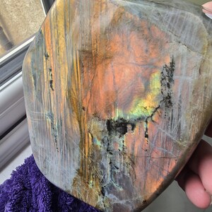 Puede incluir: Una losa de piedra labradorita pulida que presenta una superficie lisa y plana con destellos iridiscentes de color naranja, amarillo y azul. La piedra muestra fracturas internas naturales e inclusiones minerales oscuras en su base gris&aacute;cea.