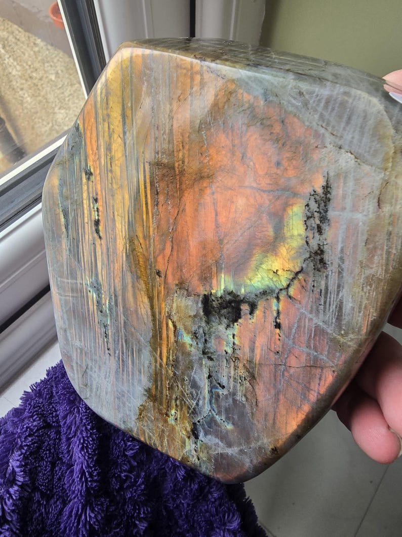 Puede incluir: Una losa de piedra labradorita pulida que presenta una superficie lisa y plana con destellos iridiscentes de color naranja, amarillo y azul. La piedra muestra fracturas internas naturales e inclusiones minerales oscuras en su base gris&aacute;cea.