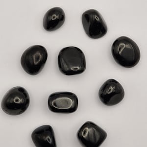 May include: A collection of nine smooth, polished black stones arranged in a scattered pattern. Each stone features a glossy, reflective surface with rounded edges and irregular, organic shapes, showcasing a deep, solid black colour throughout.