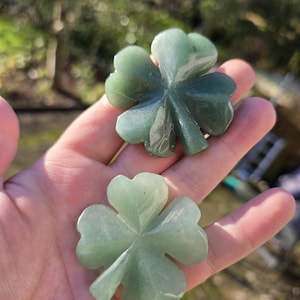 Pode incluir: Dois trevos de quatro folhas de aventurina verde. As folhas do trevo são esculpidas com veios detalhados. Os trevos são segurados em uma mão, com um fundo desfocado ao ar livre.