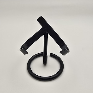 Puede incluir: Un soporte de exhibición de metal negro con una base circular y un soporte vertical central. Dos brazos curvos se extienden desde la parte superior, creando una forma de cuna. El soporte está diseñado para sostener un artículo, probablemente un plato o un pequeño objeto decorativo.