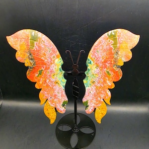 Puede incluir: Una escultura decorativa de mariposa con dos grandes alas de piedra tallada que presentan patrones vibrantes en naranja, amarillo, rosa y verde. Las alas están montadas sobre un soporte de metal negro con una base circular y un eje central.