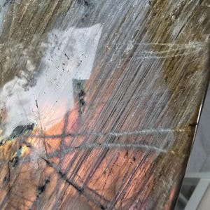 Puede incluir: Una losa pulida de piedra labradorita con una superficie lisa y reflectante con inclusiones naturales. El mineral muestra un espectro de colores iridiscentes, incluyendo tonos cobre profundo, bronce y gris apagado con estr&iacute;as lineales visibles.