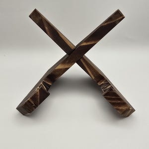 May include: A dark brown wooden book stand. The stand is made from two pieces of wood that cross each other, creating an X shape. The wood grain is visible, giving it a textured appearance. It is designed to hold books or other reading materials.