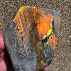 Puede incluir: Una losa de piedra labradorita pulida con una forma pentagonal irregular y lisa. La superficie muestra una labradorescencia vibrante con intensos destellos naranjas, dorados y amarillos sobre una base mineral gris oscuro semitransl&uacute;cida.