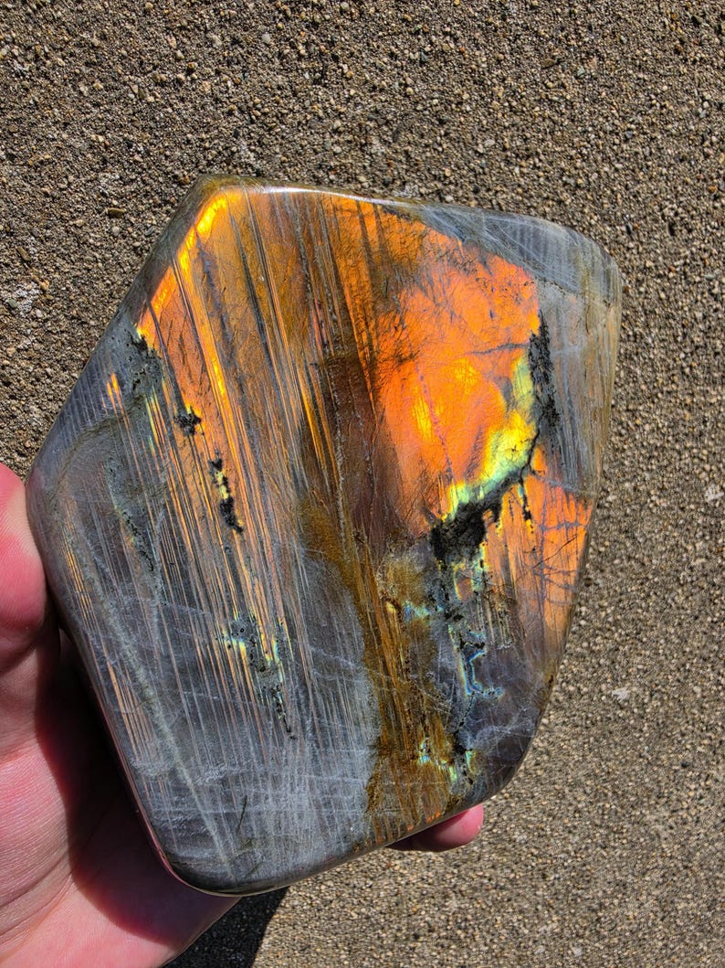 Puede incluir: Una losa de piedra labradorita pulida con una forma pentagonal irregular y lisa. La superficie muestra una labradorescencia vibrante con intensos destellos naranjas, dorados y amarillos sobre una base mineral gris oscuro semitransl&uacute;cida.