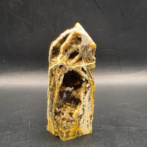 May include: A vertical, tower-shaped mineral specimen featuring a rough, textured exterior in shades of mustard yellow, brown, and cream. The front displays a deep, hollowed-out cavity filled with small, rounded, amber-coloured crystal formations.