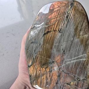 Puede incluir: Una piedra de labradorita pulida con una forma suave y redondeada. La superficie muestra una mezcla de tonos grises, marrones y naranja dorado con fracturas internas visibles y estr&iacute;as naturales que crean un aspecto iridiscente y reflectante.