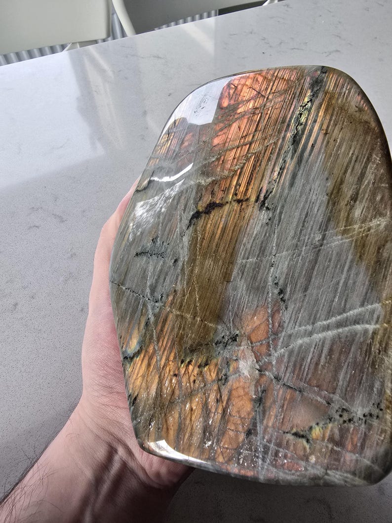 Puede incluir: Una piedra de labradorita pulida con una forma suave y redondeada. La superficie muestra una mezcla de tonos grises, marrones y naranja dorado con fracturas internas visibles y estr&iacute;as naturales que crean un aspecto iridiscente y reflectante.