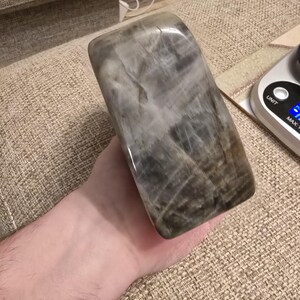 Puede incluir: Un bloque rectangular pulido de piedra gris y marr&oacute;n con vetas internas visibles y una superficie lisa y reflectante. La piedra presenta un aspecto jaspeado con varios tonos de carb&oacute;n, topo y blanco, mostrando una textura cristalina natural en toda la pieza.
