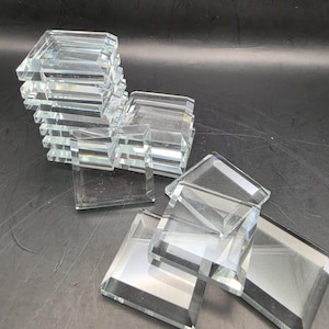 Pode incluir: Uma coleção de azulejos de vidro quadrados e transparentes com bordas chanfradas. O conjunto inclui uma pilha vertical de várias peças e vários azulejos individuais dispostos de forma dispersa, exibindo as suas superfícies transparentes e refletoras.