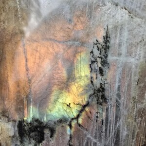 Puede incluir: Una losa de piedra preciosa labradorita pulida que presenta una superficie lisa y reflectante con destellos iridiscentes de luz amarilla, naranja y azul. La piedra muestra inclusiones minerales naturales grises y marrones, incluyendo patrones dendr&iacute;ticos oscuros.