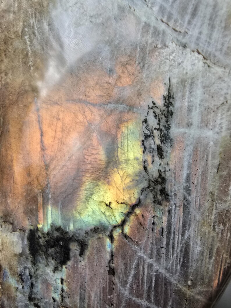 Puede incluir: Una losa de piedra preciosa labradorita pulida que presenta una superficie lisa y reflectante con destellos iridiscentes de luz amarilla, naranja y azul. La piedra muestra inclusiones minerales naturales grises y marrones, incluyendo patrones dendr&iacute;ticos oscuros.