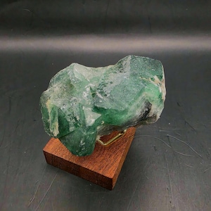 Può includere: Un grande esemplare di cristallo di fluorite verde grezzo. Il cristallo è montato su una piccola base rettangolare in legno con un supporto in ottone. La fluorite ha un aspetto naturale, non lucido, con varie tonalità di verde.