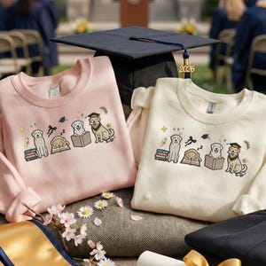 May include: Two crewneck sweatshirts, one light pink and one cream, featuring an embroidered design of graduating dogs with books and graduation caps. A black graduation cap with a gold 2026 charm is in the background.