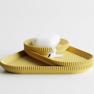 May include: Two yellow, geometric trays with a ribbed edge design. One tray holds a white charging case, and the other holds a white earbud. The trays are stacked on top of each other.