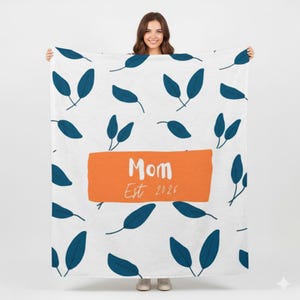 Puede incluir: Manta blanca con un estampado de hojas azules. Un rectángulo naranja en el centro tiene la palabra "Mom" y "Est 2026" impresas en blanco. La manta es sostenida por una persona.