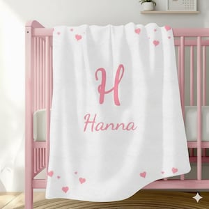 Puede incluir: Manta de bebé blanca con corazones rosas y el nombre "Hanna" en escritura rosa. Una gran letra "H" rosa está centrada encima del nombre. La manta está sobre una cuna rosa.