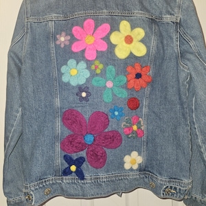 May include: A denim jacket with a variety of colorful, felt flowers appliqued on the back. The flowers are in shades of pink, yellow, blue, green, and purple. The jacket has a classic design with button closures and pockets.