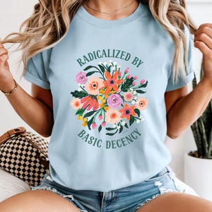 Puede incluir: Camiseta azul claro con el texto "RADICALIZED BY BASIC DECENCY" y un diseño floral en tonos rosas, naranjas y amarillos. La camiseta es de manga corta y está hecha de un material suave.