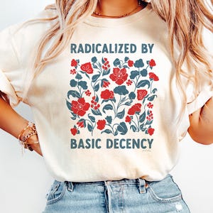 Puede incluir: Camiseta color crema con el texto "RADICALIZED BY BASIC DECENCY" encima y debajo de un diseño floral de flores rojas y hojas azules. La camiseta se combina con vaqueros de mezclilla azul claro.