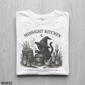 May include: A white t-shirt featuring a black graphic of a cat wearing a witch hat, stirring a cauldron. The text reads MIDNIGHT KITCHEN, RECIPES for MAGIC, MOON DUST, RAT WINGS, WORM WOOD, NIGHT SHADE, and STIRRING UP MAGIC ONE SPELL AT A TIME.