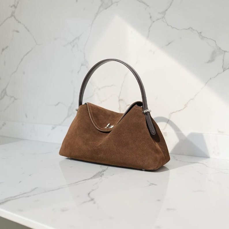 K&ouml;nnte beinhalten: Eine dunkelbraune Wildlederhandtasche mit einer strukturierten, trapezf&ouml;rmigen Silhouette. Die Tasche verf&uuml;gt &uuml;ber einen einzelnen dunkelbraunen Ledergriff, der mit silberfarbenen Metallbeschl&auml;gen befestigt ist, und einen silberfarbenen Metallverschluss.