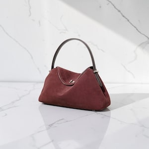 K&ouml;nnte beinhalten: Eine tiefburgunderrote Wildlederhandtasche mit strukturierter Silhouette, einem einzelnen dunkelbraunen Ledergriff und einem silberfarbenen Metalldrehverschluss, der mittig auf der Frontklappe angebracht ist. Die Tasche hat eine glatte, matte Textur.