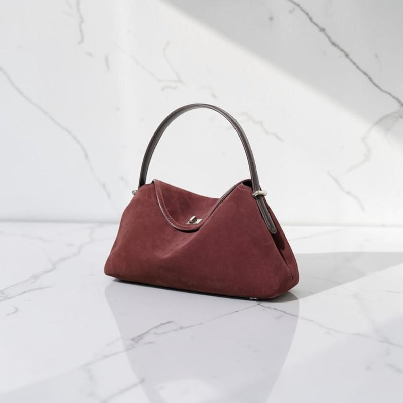 K&ouml;nnte beinhalten: Eine tiefburgunderrote Wildlederhandtasche mit strukturierter Silhouette, einem einzelnen dunkelbraunen Ledergriff und einem silberfarbenen Metalldrehverschluss, der mittig auf der Frontklappe angebracht ist. Die Tasche hat eine glatte, matte Textur.