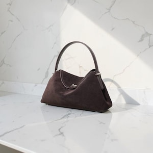 K&ouml;nnte beinhalten: Eine dunkelbraune Wildlederhandtasche mit einem einzelnen, gebogenen Tragegriff und einem silberfarbenen Metallverschluss. Die Tasche hat eine strukturierte, l&auml;ngliche Basis und eine weiche, matte Textur mit silbernen Beschl&auml;gen am Griff.