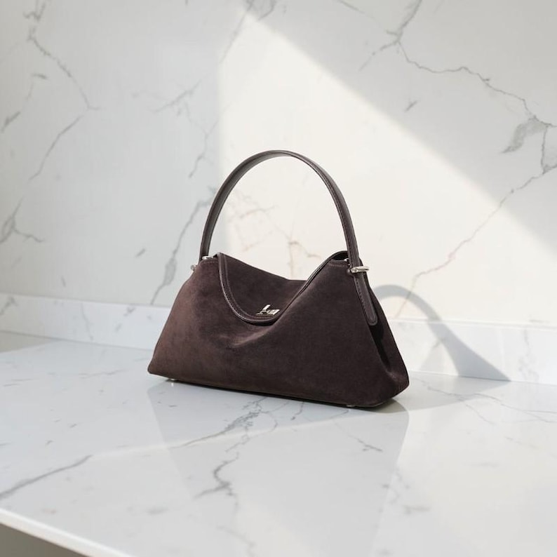 K&ouml;nnte beinhalten: Eine dunkelbraune Wildlederhandtasche mit einem einzelnen, gebogenen Tragegriff und einem silberfarbenen Metallverschluss. Die Tasche hat eine strukturierte, l&auml;ngliche Basis und eine weiche, matte Textur mit silbernen Beschl&auml;gen am Griff.