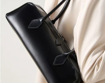 Könnte beinhalten: Eine elegante, rechteckige schwarze Handtasche mit glatter, polierter Oberfläche. Die Tasche verfügt über zwei schmale Tragegriffe, die mit spitzen, rautenförmigen Laschen befestigt sind, sowie einen Reißverschluss mit passendem schwarzem Zipper.