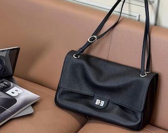 Könnte beinhalten: Eine schwarze Lederschultertasche mit genarbter Textur, ausgestattet mit einer rechteckigen Klappe, einem silberfarbenen Drehverschluss aus Metall und verstellbaren Schulterriemen mit silbernen Beschlägen. Daneben liegen eine schwarze Sonnenbrille mit der Aufschrift CHANEL.